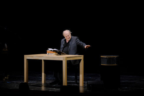 Klaus Maria Brandauer sitzt ein Buch vorlesend an einem Tisch