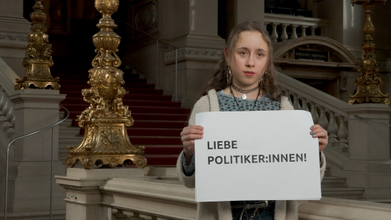 Ausschnitt aus dem Video das im Rahmen des Welttag des Theaters für junges Publikum gedreht wurde. Es zeigt ein junges Mädchen mit einem Plakat auf dem steht: Liebe Politiker*innen