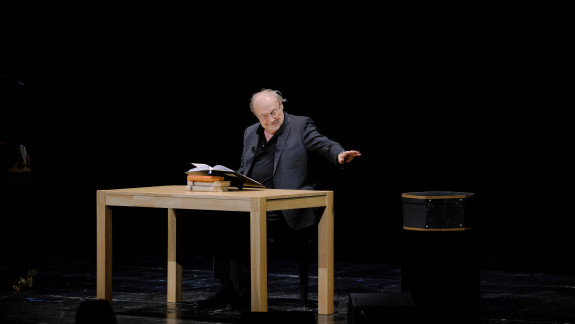 Klaus Maria Brandauer sitzt ein Buch vorlesend an einem Tisch